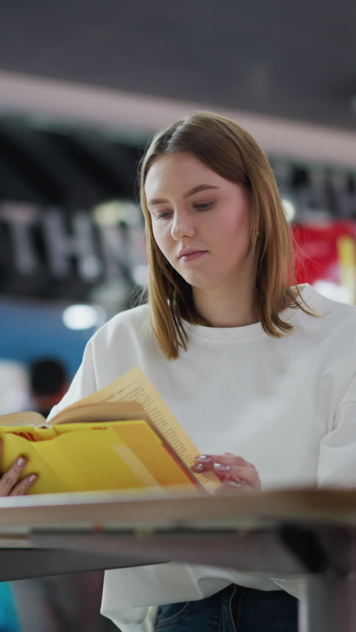 Young woman reading her book with coffee cup on table in lively indoor mall setting, soft blur background features vibrant colors, shopping bags nearby and other shoppers