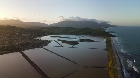 Aerial-view-during-sunset-of-salt-flats-near-slums,-with-green-lush-mountains-and-waves-splashing-across-the-coast-in-Isla-de-Margarita,-Venezuela