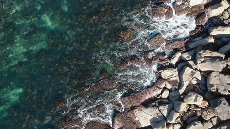 Top-down-birds-eye-view-drone-shot-of-waves-crashing-into-jagged-rocks-off-the-coastline-of-Australia