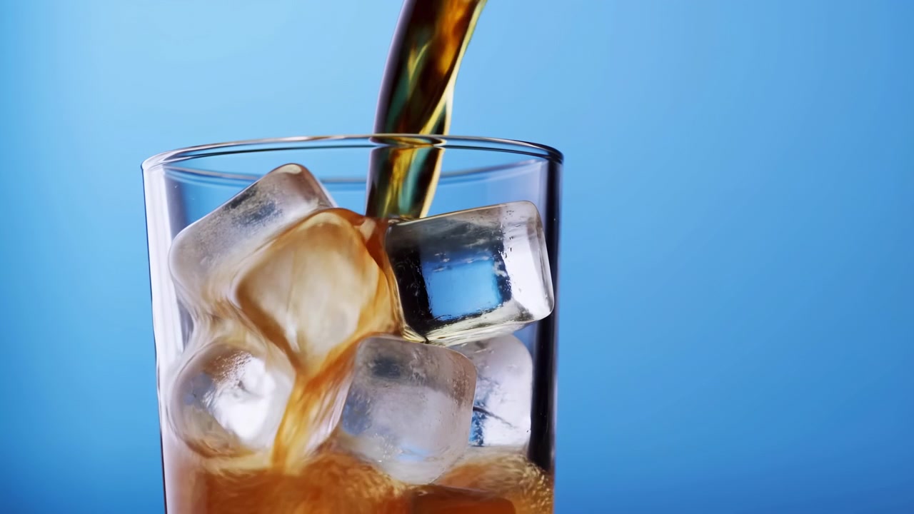 Versamento progressivo di una bevanda scura gassata in un bicchiere pieno di ghiaccio su uno sfondo sfumato blu.
