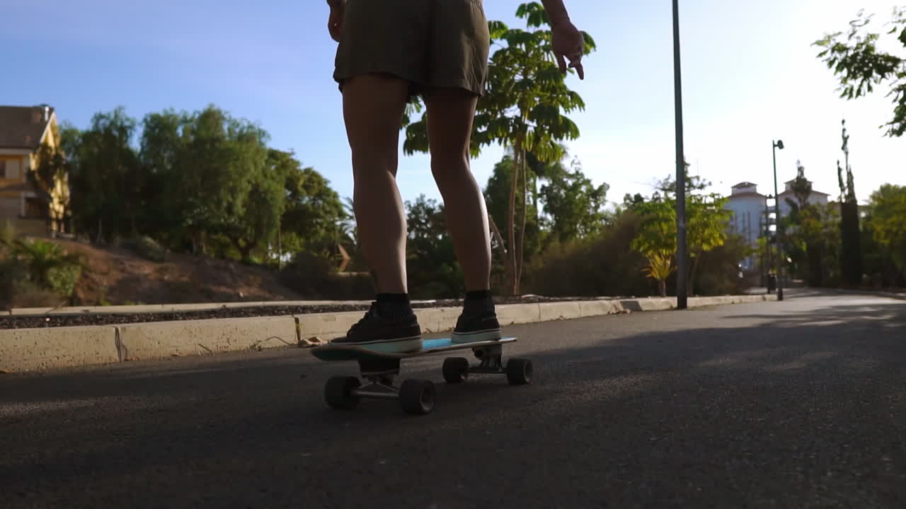 Against the backdrop of sunset hues, a woman on a skateboard glides along the path in the park, grinning, with palm trees and sandy surroundings. Happy individuals living healthily