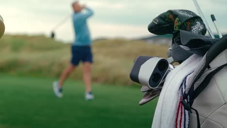 male golfer takes drive off of tee box in slow motion blurry in background with golf bag full of clubs and range finder are in focus in foreground on beautiful green golf course