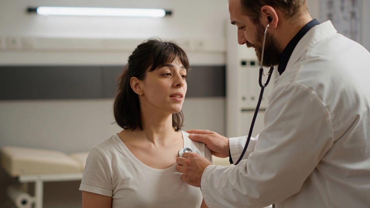 Stetoskopu yerleştiren doktor, klinikte beyaz tişörtlü kadın hastanın akciğerlerini dinleyerek değerlendiriyor.
