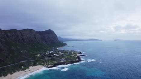 An-aerial-perspective-of-North-Shore-Oahu-reveals-the-majestic-cliffs-meeting-the-vibrant-blue-ocean,-with-a-serene-beach-and-a-small-coastal-community-nestled-at-the-base