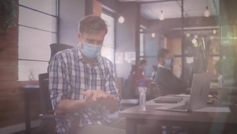 Animation-of-glowing-spots-over-man-in-office-sanitizing-his-hands-wearing-face-mask