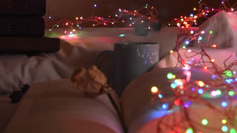 cozy background of an open book on a bed, next to a mug and surrounded by colorful christmas lights