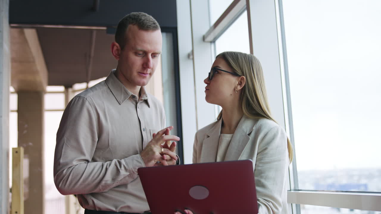 Office coworkers both look into the laptop standing at the window. Woman holds a computer and tells something to the man who replies afterwards.