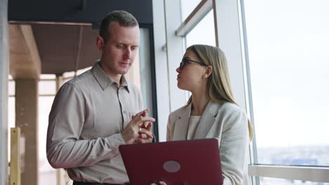 office coworkers both look into the laptop standing at the window. woman holds a computer and tells something to the man who replies afterwards.