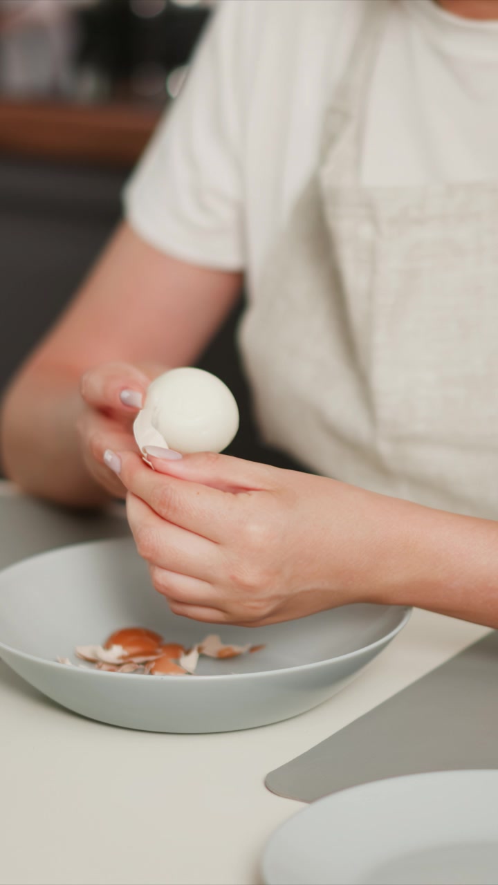 Woman carefully peels hard boiled egg holding it in one hand while removing shell with other. process is shown up close as woman places peeled shells into small bowl on clean countertop