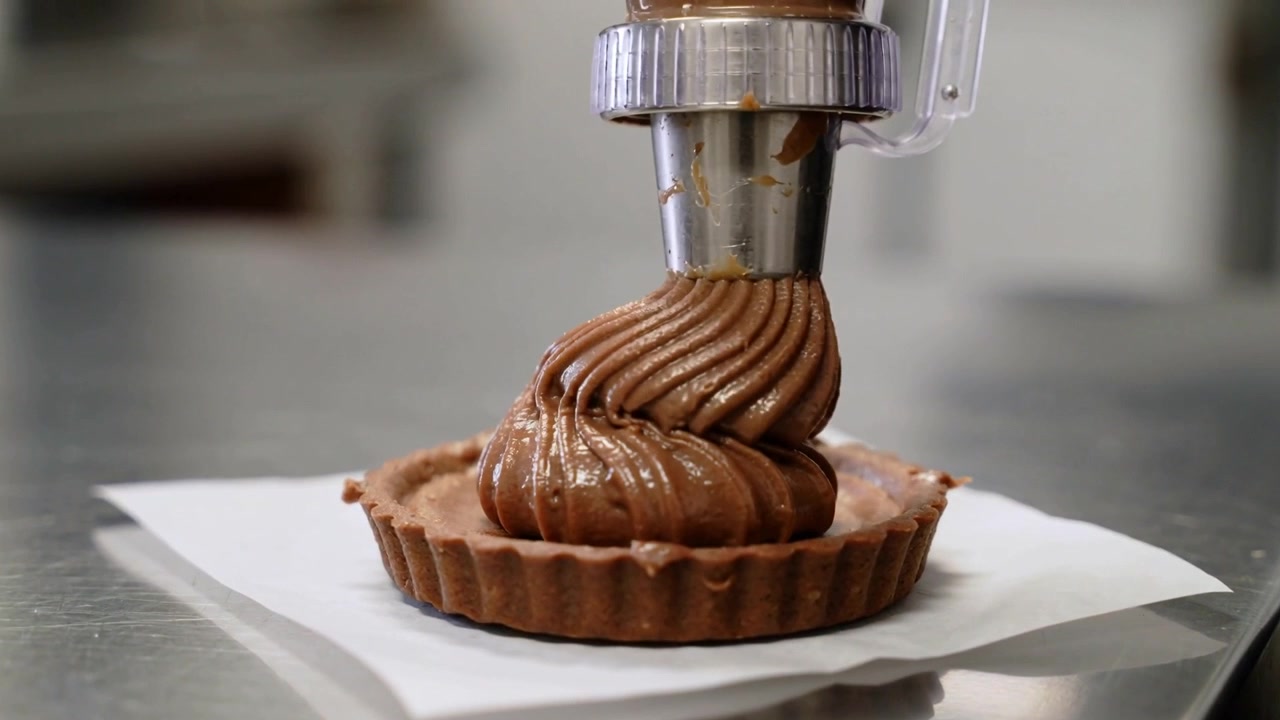 Piping nozzle descending to touch base, extruding chocolate ribbon swirl to fill shell in bakery