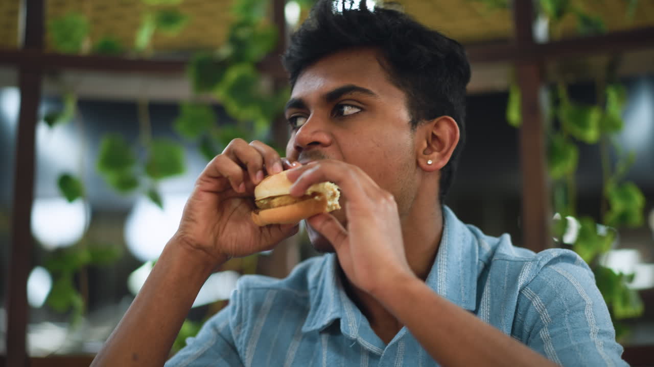 Student wearing earring eating burger while looking off camera, closeup showing hand holding sandwich, mouth mid bite, casual indoor setting, expression of enjoyment, background softly out of focus