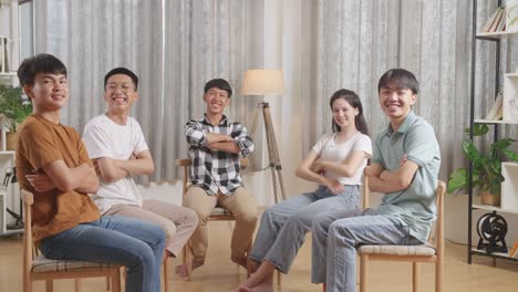 asian teen group sitting in chairs forming a circle discussing at home. smiling and showing arms crossed gesture to camera