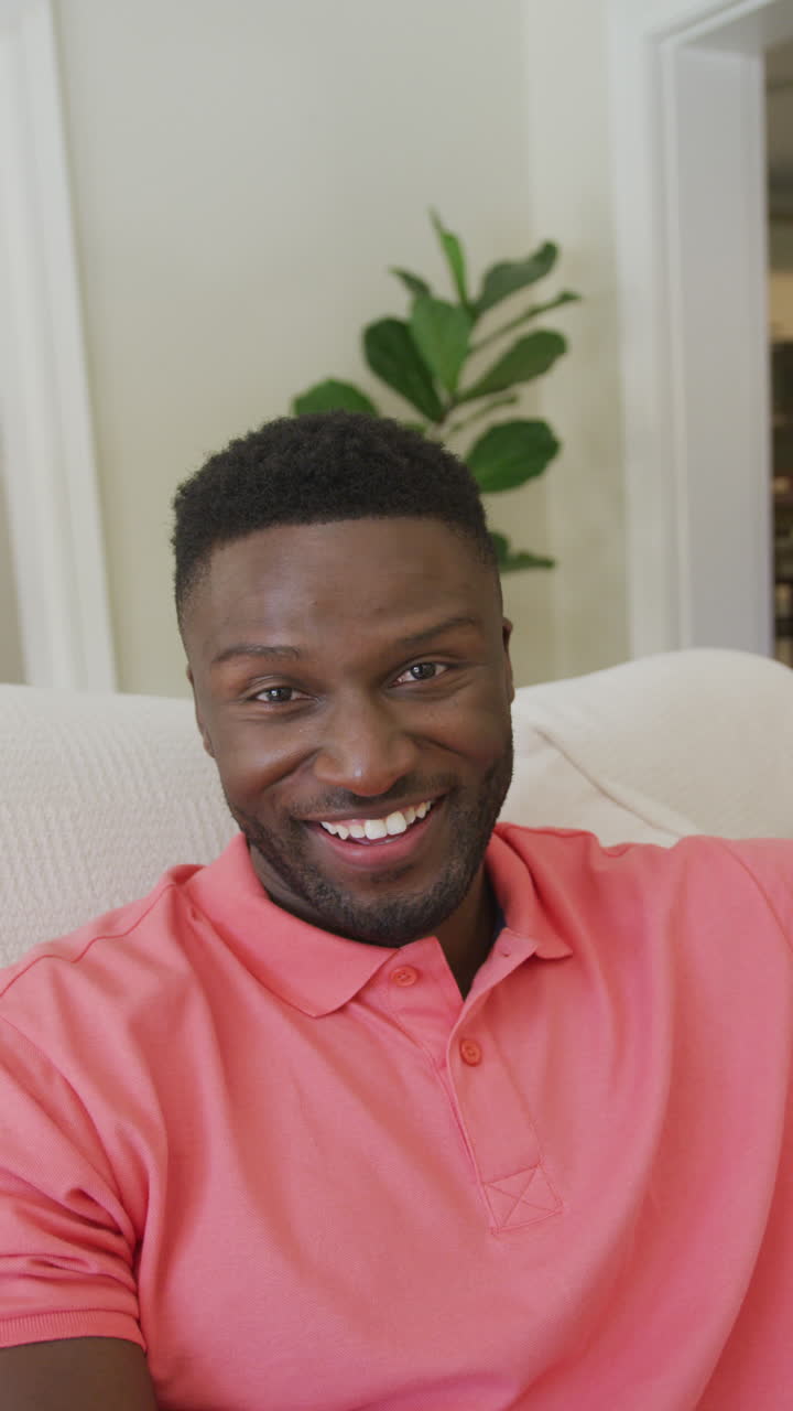 retrato vertical de video de un feliz hombre afroamericano sentado en el sofá sonriendo en la sala de estar