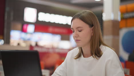 mujer concentrada trabajando remotamente en un restaurante usando una computadora portátil, deteniéndose brevemente para mirar hacia otro lado antes de reanudar el trabajo, fondo borroso vibrante