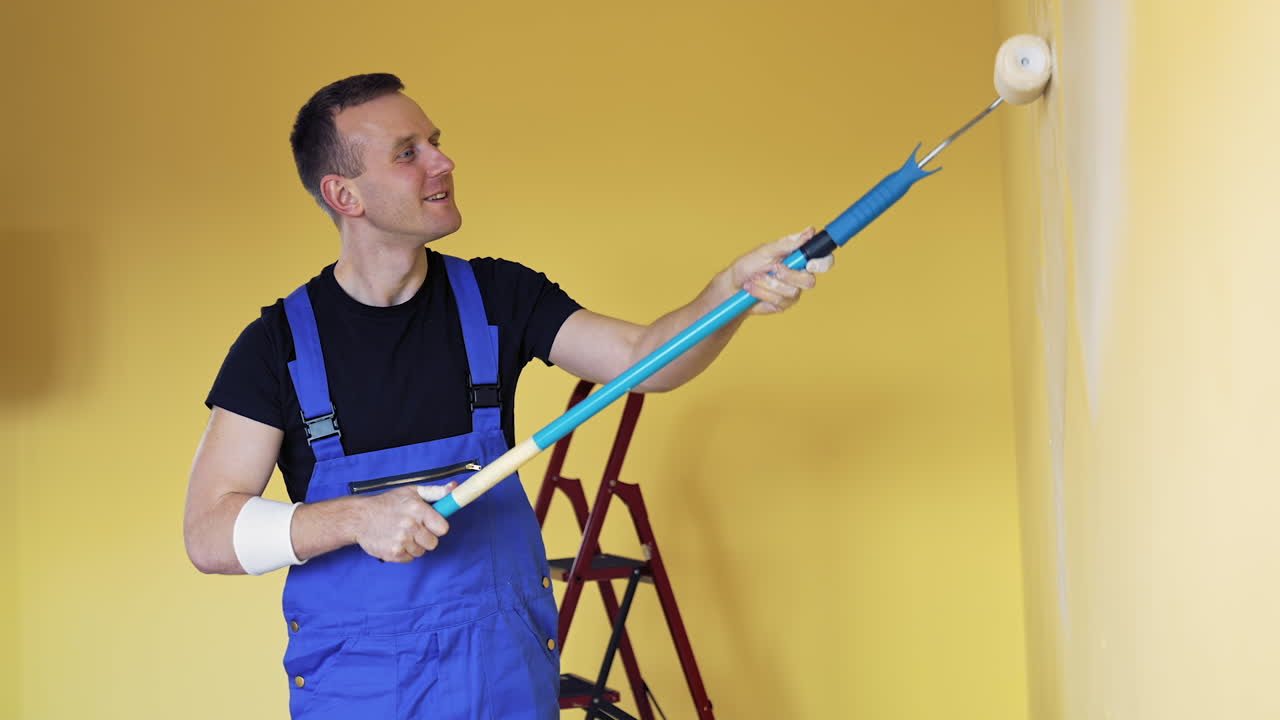 Young man doing renovation. Portrait of a painter worker doing makeover in the room. Worker in overalls paints wall with a roller brush.