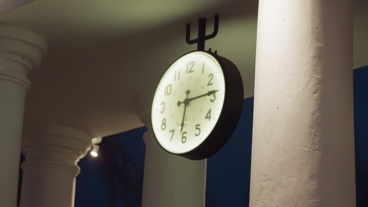 Large round wall clock with black frame and white illuminated face hanging under roof structure with classical white pillars, captured at night with soft lighting casting shadows on smooth surfaces