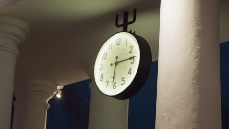 large round wall clock with black frame and white illuminated face hanging under roof structure with classical white pillars, captured at night with soft lighting casting shadows on smooth surfaces
