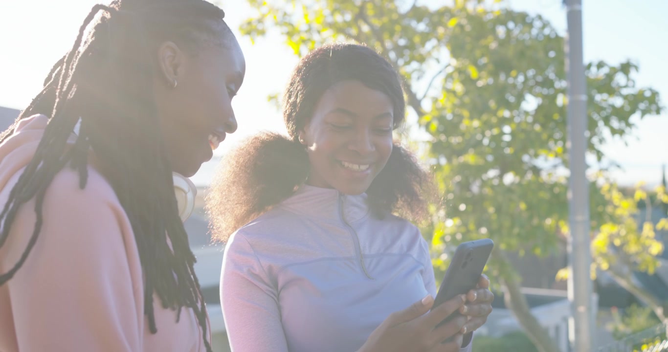 African female friends with hoodie leaning on street, right woman tapping phone showing funny text
