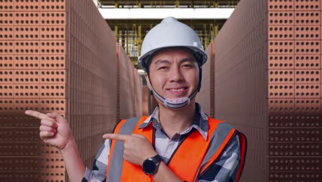close up of asian male engineer with safety helmet smiling and pointing to side while standing with red brick packed in stacks are stored