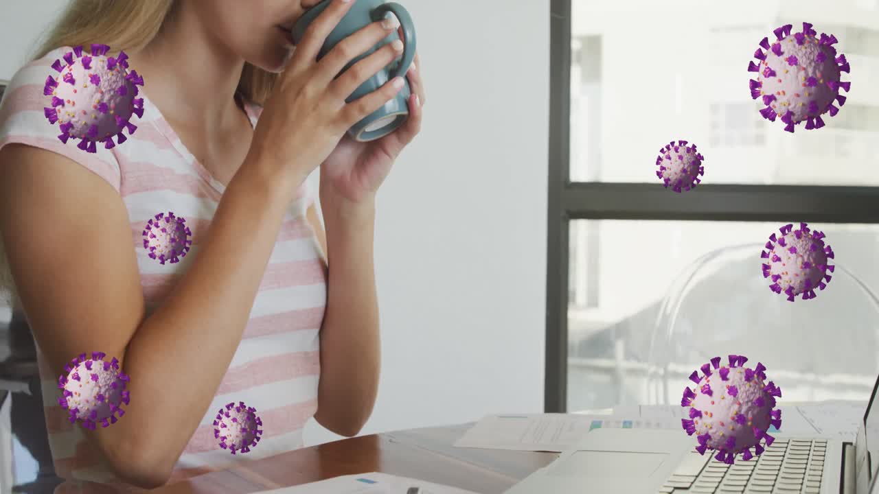 Animazione di cellule di coronavirus su una giovane donna bianca che beve caffè mentre lavora alla scrivania.