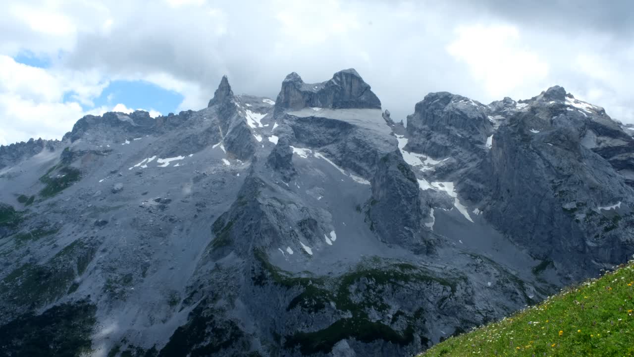 Majestic gray mountains with steep cliffs and snow traces rise above lush valleys, captured beneath a cloudy sky in a stunning aerial or elevated view.