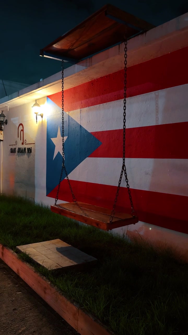 San Juan, Porto Rico, 4 agosto 2025: Altalena di legno davanti a un murale della bandiera portoricana. Un'altalena di legno vuota è appesa a catene davanti a un muro dipinto con una grande bandiera portoricana rossa, bianca e blu di notte