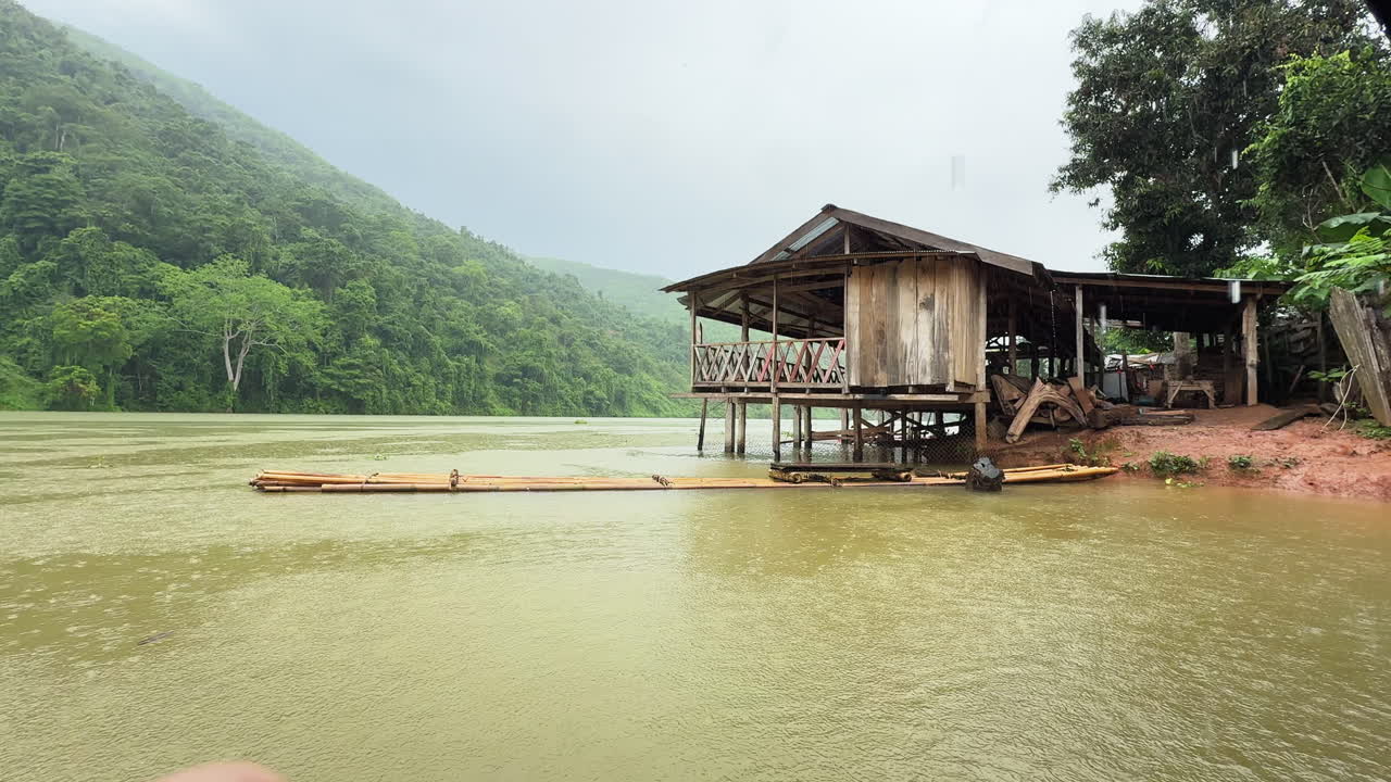 Valle del fiume Nam Ou, piovosa, in Laos, con una rustica casa di legno su palafitte sulla riva fangosa e una semplice zattera di bambù che galleggia accanto alle colline boscose
