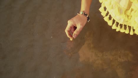 Mano-De-Mujer-Con-Una-Pulsera-Marrón-Y-Blanca-Recogiendo-Una-Concha-Marina-Del-Agua-En-La-Playa