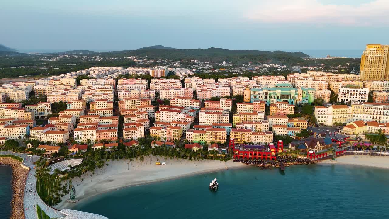 Aerial view of the sunset over Phu Quoc Island, Vietnam, highlighting a replica Italian-style town by the beach. The colorful buildings along the coastline contrast with the serene waters and hills.