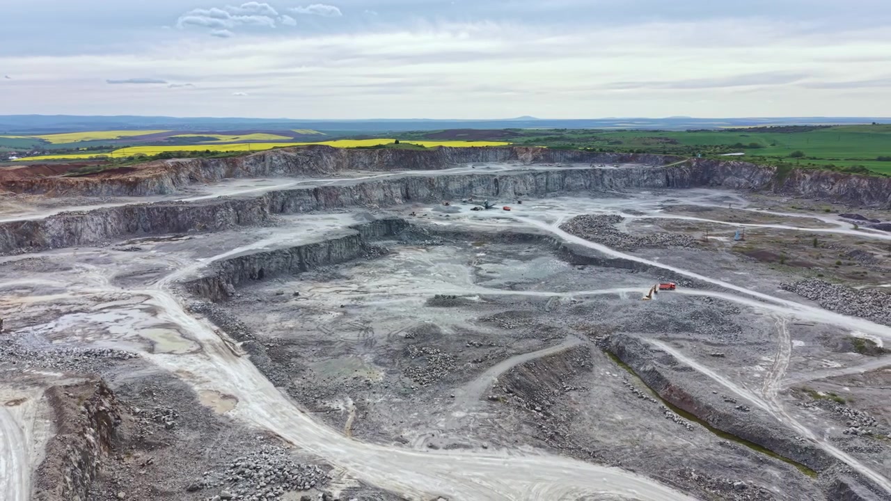 Workers extract stone from a large quarry in the countryside while machinery operates and clouds drift overhead in a clear sky