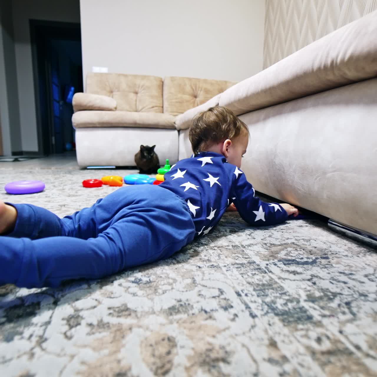 Toddler wearing blue clothes lies on the floor looking under the sofa. Baby pulls the phone from under the couch and then pushes back