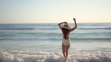 una mujer joven y saludable con cabello largo y sombrero para el sol corre hacia las olas del océano