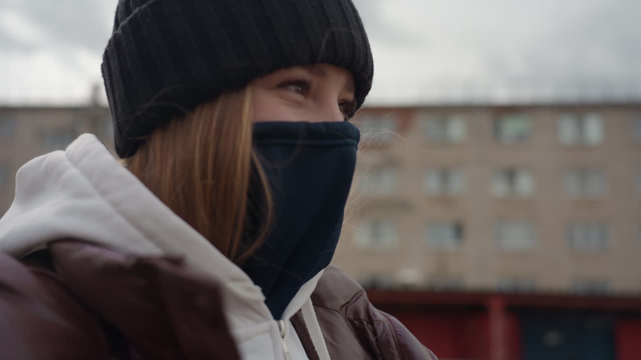 Молодая женщина в черной шапке, стоящая на улице в городском районе, в белой толстовке и коричневой кожаной куртке, в темной маске, закрывающей нижнюю часть лица, с нейтральным выражением лица наблюдает за улицей