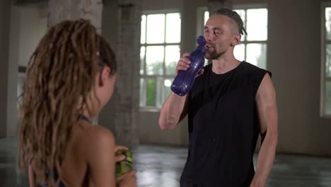 Stylish-sporty-man-and-woman-standing-in-studio-sport-club-with-windows-background-and-talking.-They-are-drinking-water-after-workout-with-color-bottles.-Both-with-stylish-dreadlocks.-Slow-motion