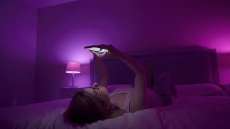 girl laying on back browses social media on smart phone late at night in bedroom.