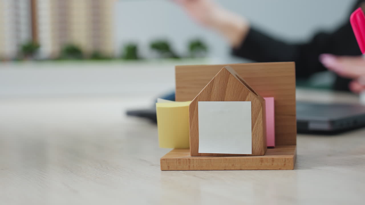 Close up of wooden desk organizer holding sticky notes on office table, while blurred professional in background gestures with pencil during project explanation