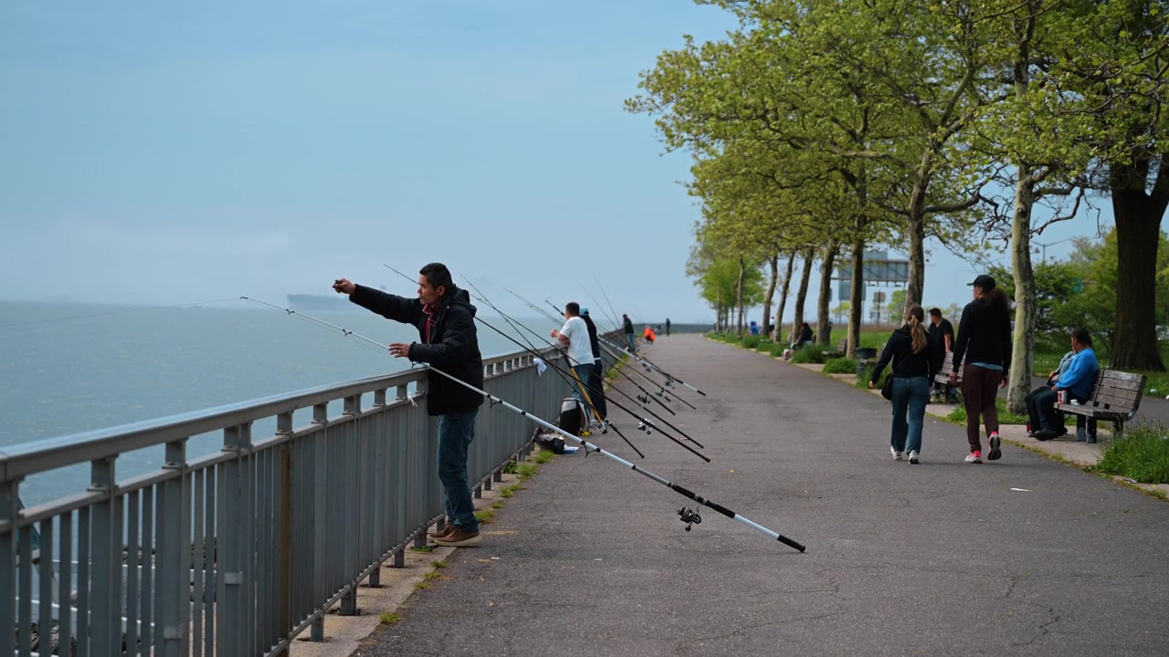 New York, USA, 1 Mei 2026: Orang memancing dengan joran di sepanjang pagar tepi air dekat pepohonan. Pemancing berdiri di jalur taman beraspal di samping air yang tenang pada siang hari.
