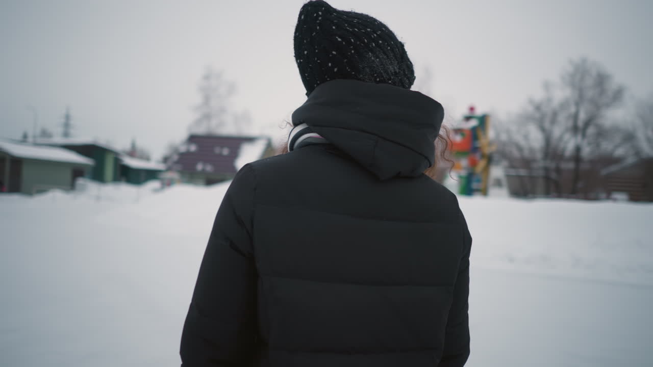 Lady skating outdoors in snowy winter park wearing black coat, knitted hat, striped scarf, and gloves, enjoying seasonal activity surrounded by snow, trees, and buildings under cold cloudy sky