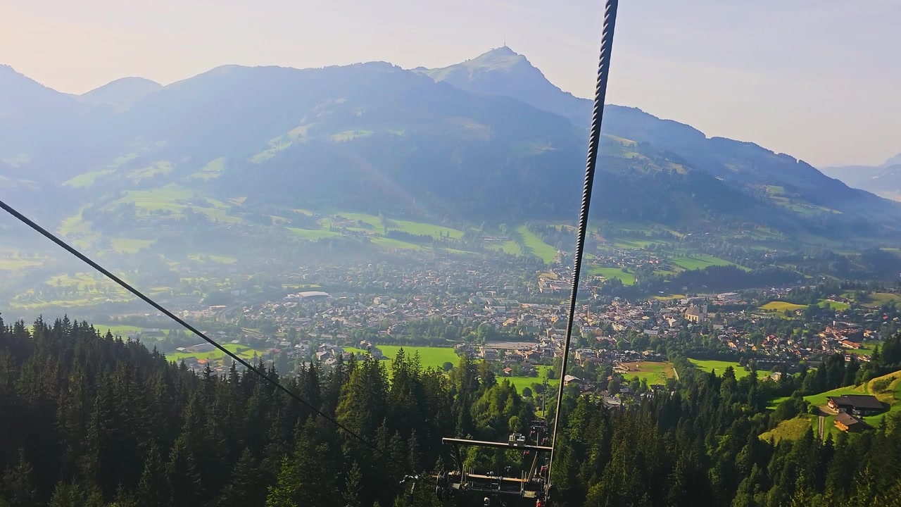 Punto di osservazione elevato su una località turistica alpina con linee di funivia, tetti di case del villaggio, pendii boscosi, pascoli verdi e un vasto paesaggio montano sotto il sole estivo per il turismo