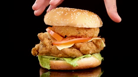 A hand gently places a sesame seed bun atop a stacked crispy chicken burger with lettuce, cheese, tomato, and onion ring against a black background. Studio lighting, close-up shot