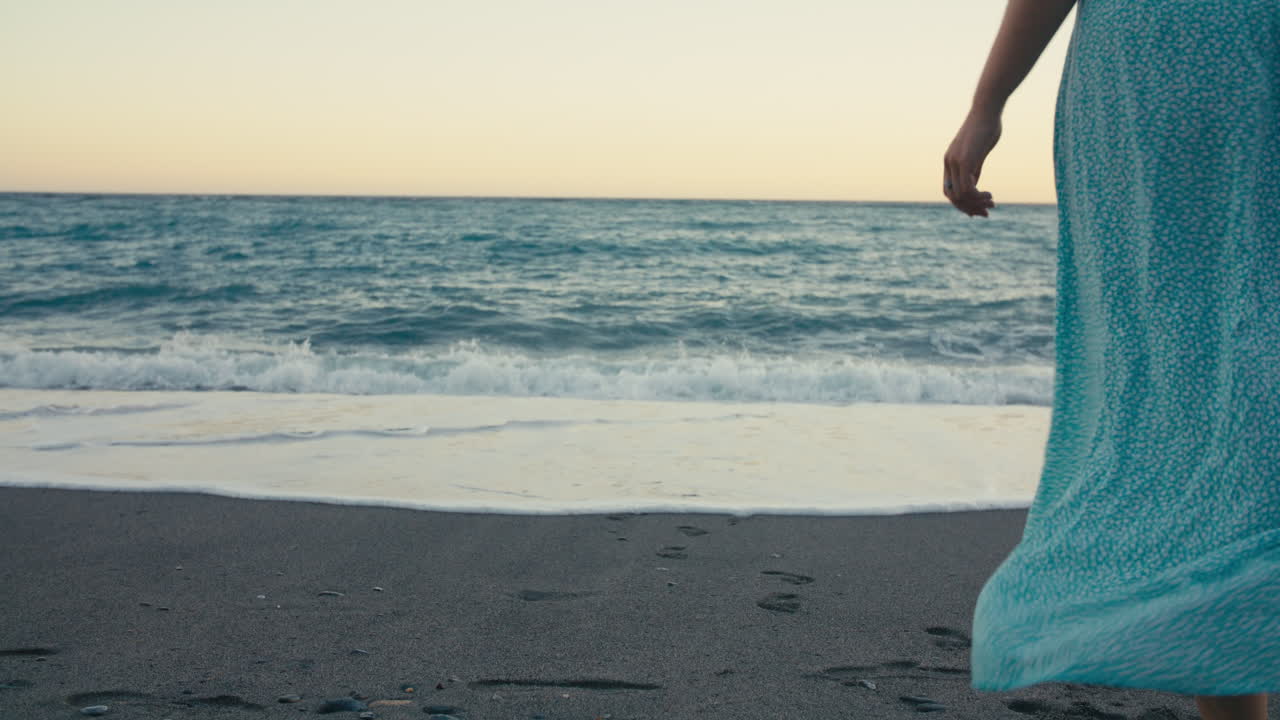 Medium rear shot of young woman walking on wet sand, her blue skirt catching the breeze, with soft evening light over the beautiful mediterranean sea