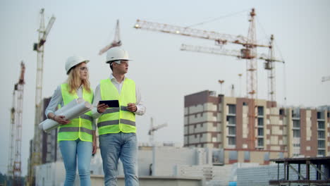 adult engineer man and architect woman uses a tablet in operation. writes a message or checks a drawing. against background is building. see neighborhood and drawings for verification of completed and planned construction works. the work of a crane