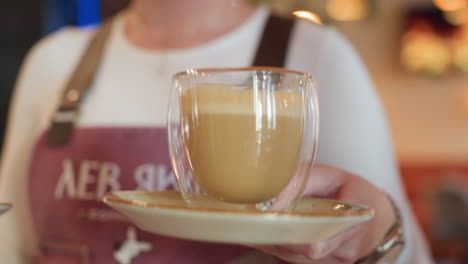 close up of bartender hand holding saucer and glass cup of frothy coffee, poised to serve customer in warm indoor coffee bar setting, with blurred barista apron and soft lighting in background