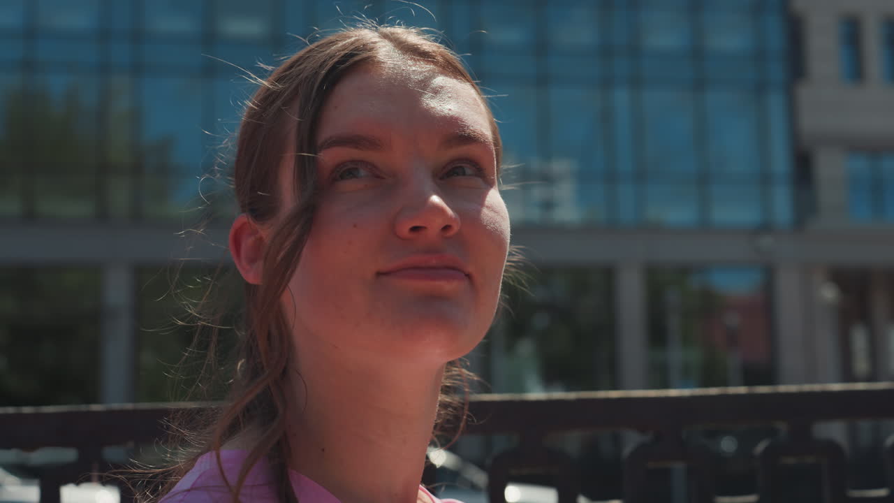 young woman on city bench sunlit, daydreaming and glancing upward, gentle breeze moves hair candid summer portrait with glass building backdrop, soft rim light on face, introspective mood, subtle