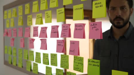 serious young caucasian man writes creative ideas on pink sticky notes on mirror to plan and organize work project, adds note with word conclusion, static, closeup