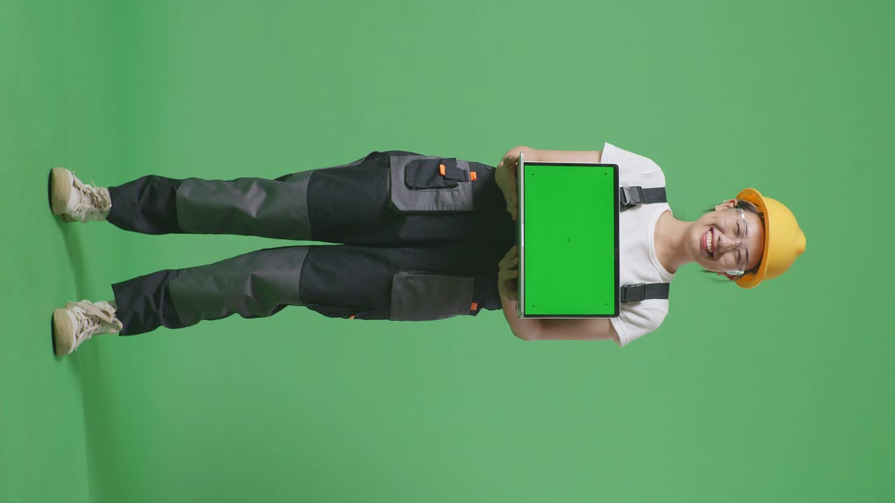 cuerpo lleno de mujer asiática trabajadora con gafas de protección y casco de seguridad sonriendo y mostrando portátil de pantalla verde a la cámara mientras está de pie en el estudio de fondo de pantalla verde