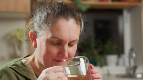 middle aged woman in green dress and white apron enjoying hot coffee from transparent glass cup with eyes closed, savoring aroma in cozy kitchen surrounded by soft lighting