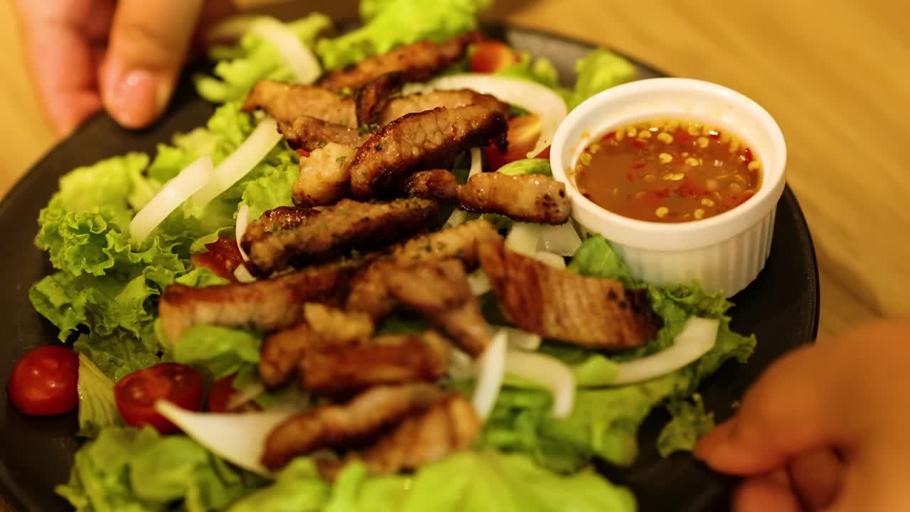 Hands carefully arrange a salad with grilled pork, lettuce, and a side of sauce on a wooden table.