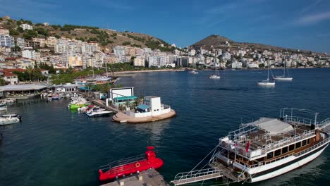 Saranda-Serenidad-Belleza-Costera-Mediterránea-Con-Un-Muelle,-Barcos-Anclados-Y-Barcos-Adornando-La-Bahía-Azul-Celeste