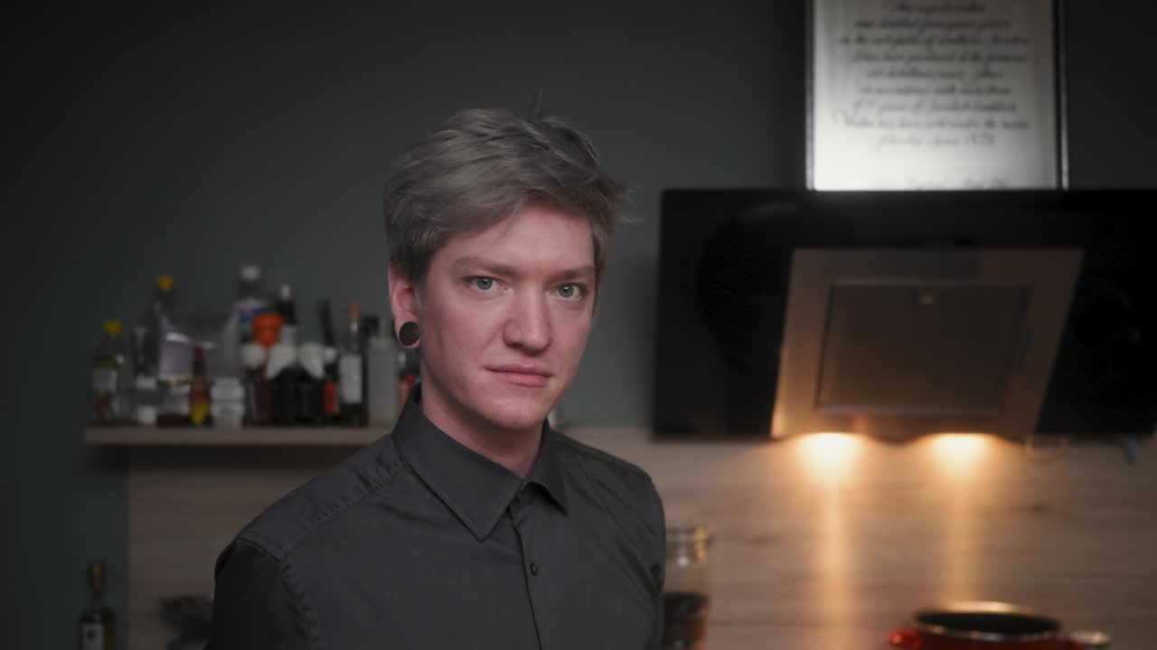 우아한 검은색 셔츠를 입은 전문적인 매력적인 남성 요리사가 붉은 고추를 물고 현대적이고 멋진 주방을 카메라에 대고 윙크하면서 느린 모